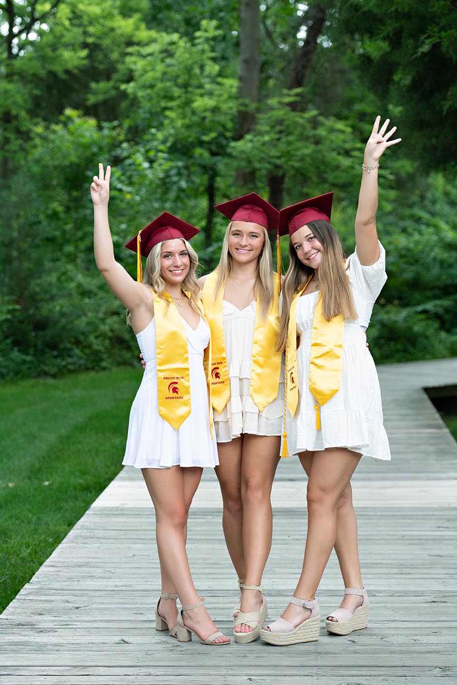 Three high school girls standing together in white dresses and graduation caps on wooden deck.