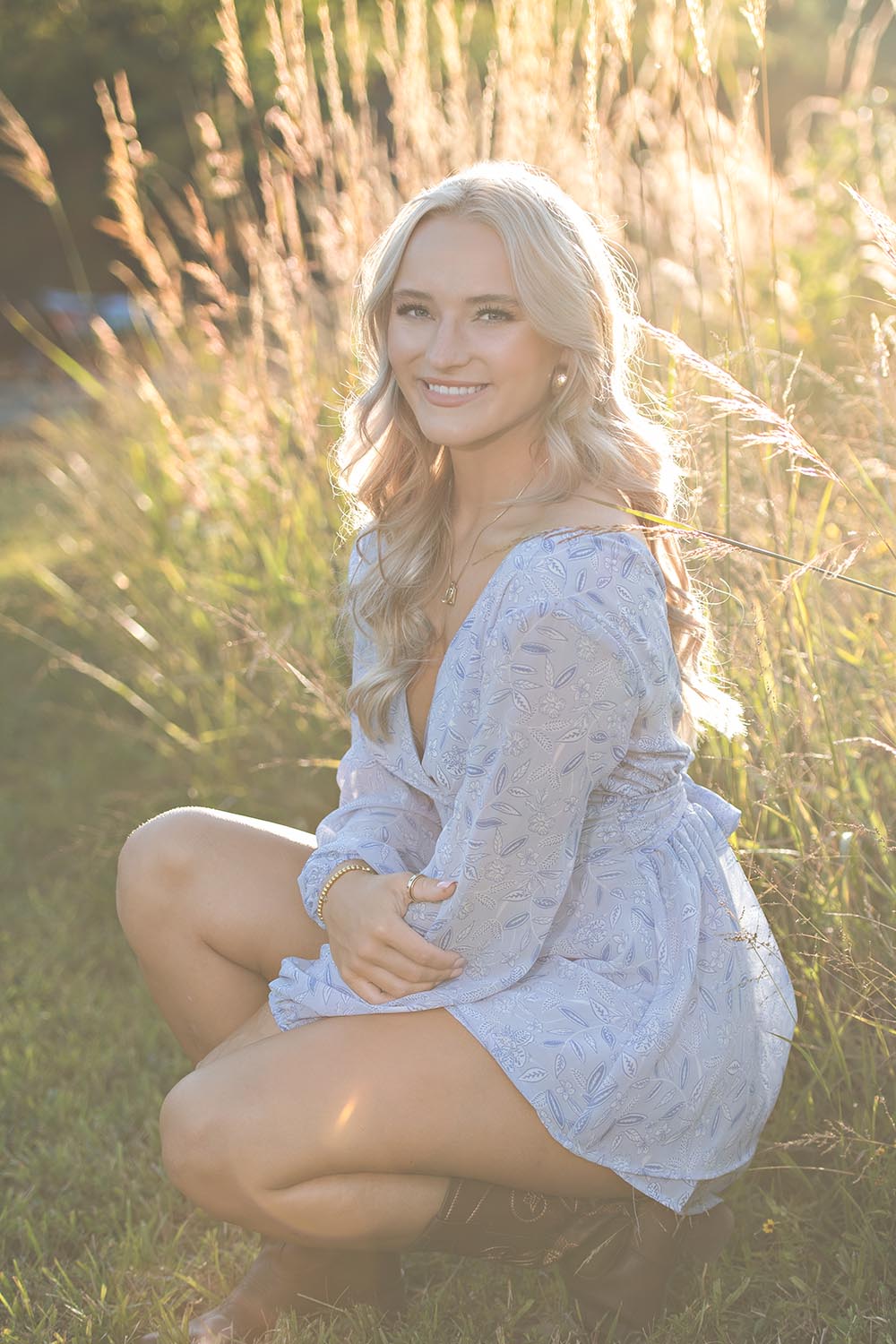 A high school senior girl kneeling in sunny grassy field.