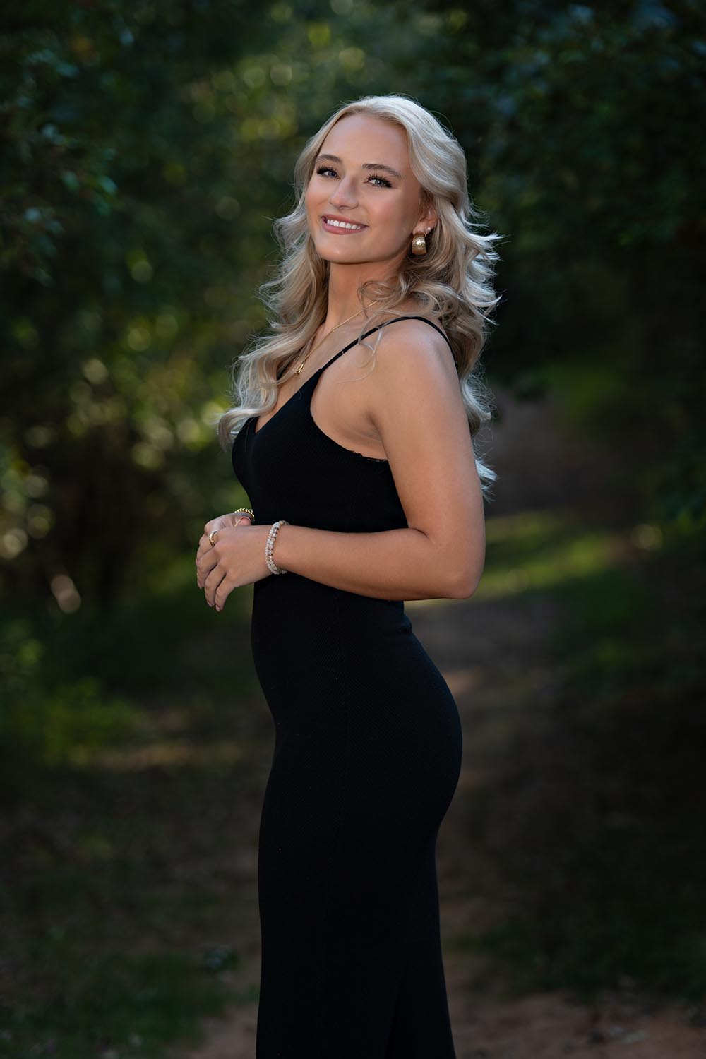 A high school senior girl posing in a black dress near wooded area.